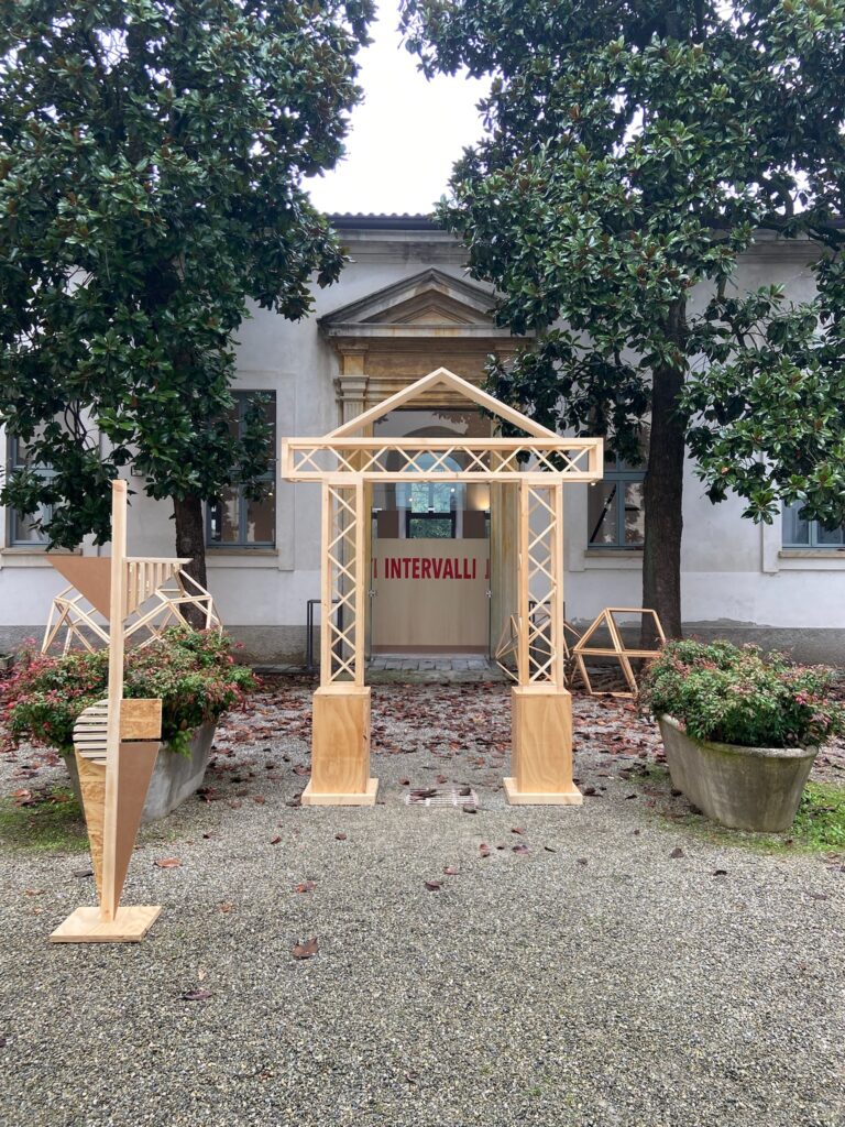 Foto dell'ingresso alla mostra con un arco in legno e alcune strutture in legno realizzate dagli studenti